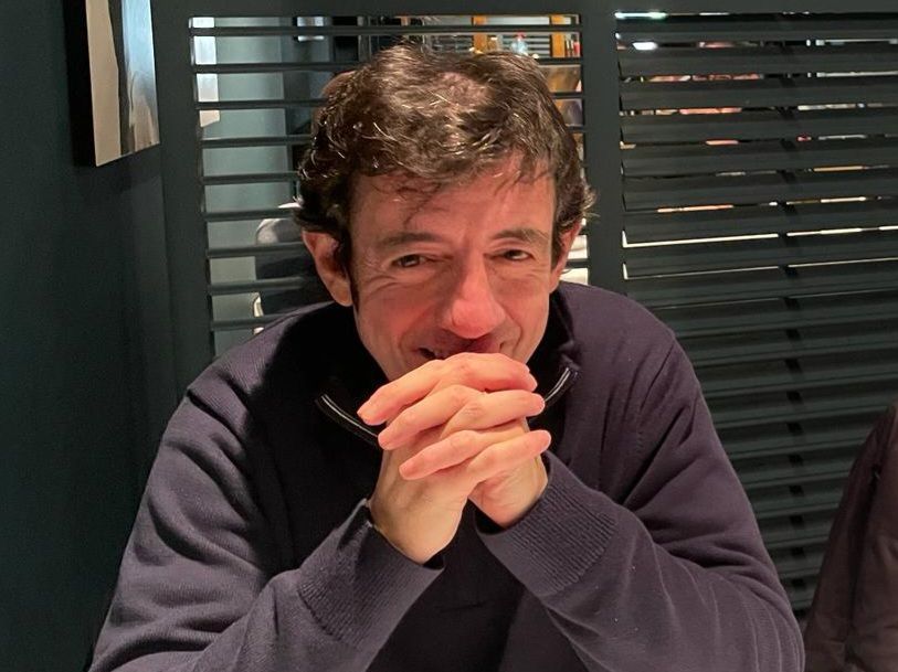 Hombre de cabello oscuro, con las manos juntas, sonriente, vestido con un suéter azul. Sentado a la mesa en un restaurante.