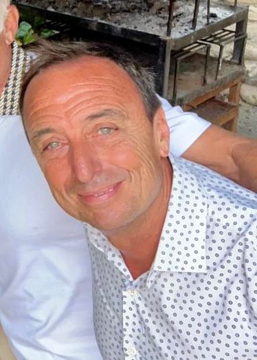 Un hombre de ojos azules y una leve sonrisa, vestido con una camisa blanca con pequeños lunares negros, posa al aire libre.