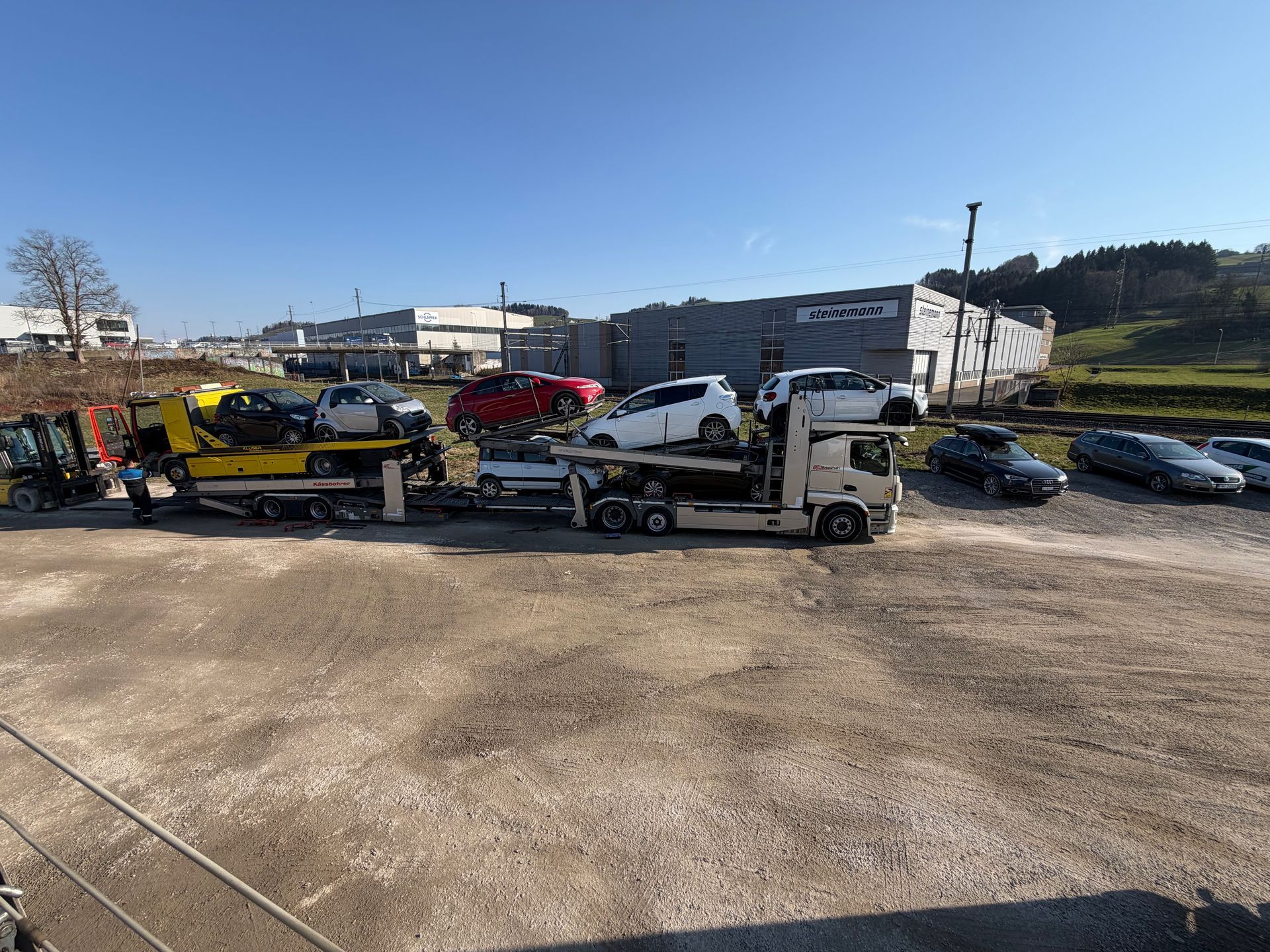 Ein Abschleppwagen verlädt an einem sonnigen Tag einen weissen Pkw auf einem Parkplatz mit mehreren anderen Fahrzeugen.