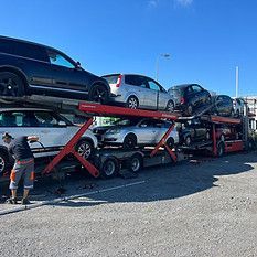 Ein beladener Autotransporter steht mit Fahrzeugen auf einem Parkplatz am Strassenrand unter strahlend blauem Himmel