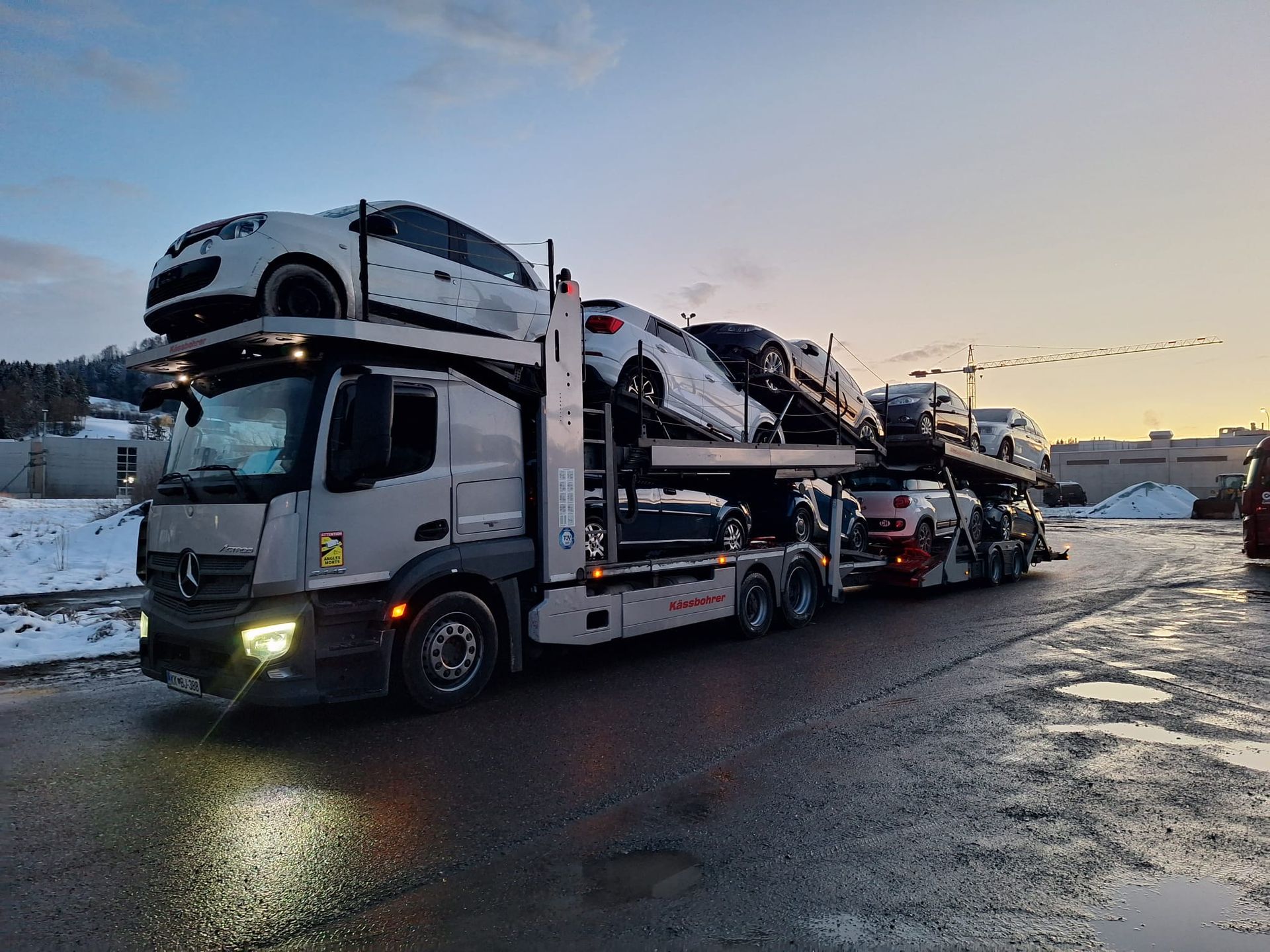 Ein mit Autos beladener Autotransporter steht in der Abenddämmerung auf nasser Strasse, umgeben von Schnee.