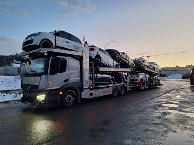 Ein mit Autos beladener Autotransporter steht in der Abenddämmerung auf nasser Strasse, umgeben von Schnee.