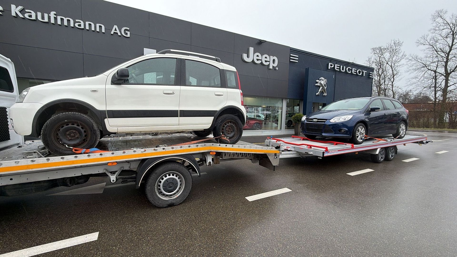 Zwei Autos auf Abschleppwagen vor einem Jeep-Händler am nassen Strassenrand.