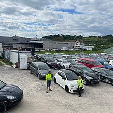 Parkplatz voller Autos, neben einem weissen Auto zwei Arbeiter in gelben Westen unter bewölktem Himmel.