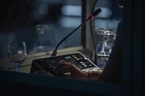 Eine Person bedient in einer schwach beleuchteten Kabine ein Bedienfeld mit einem Mikrofon, neben einem Glas und einer Wasserkaraffe.