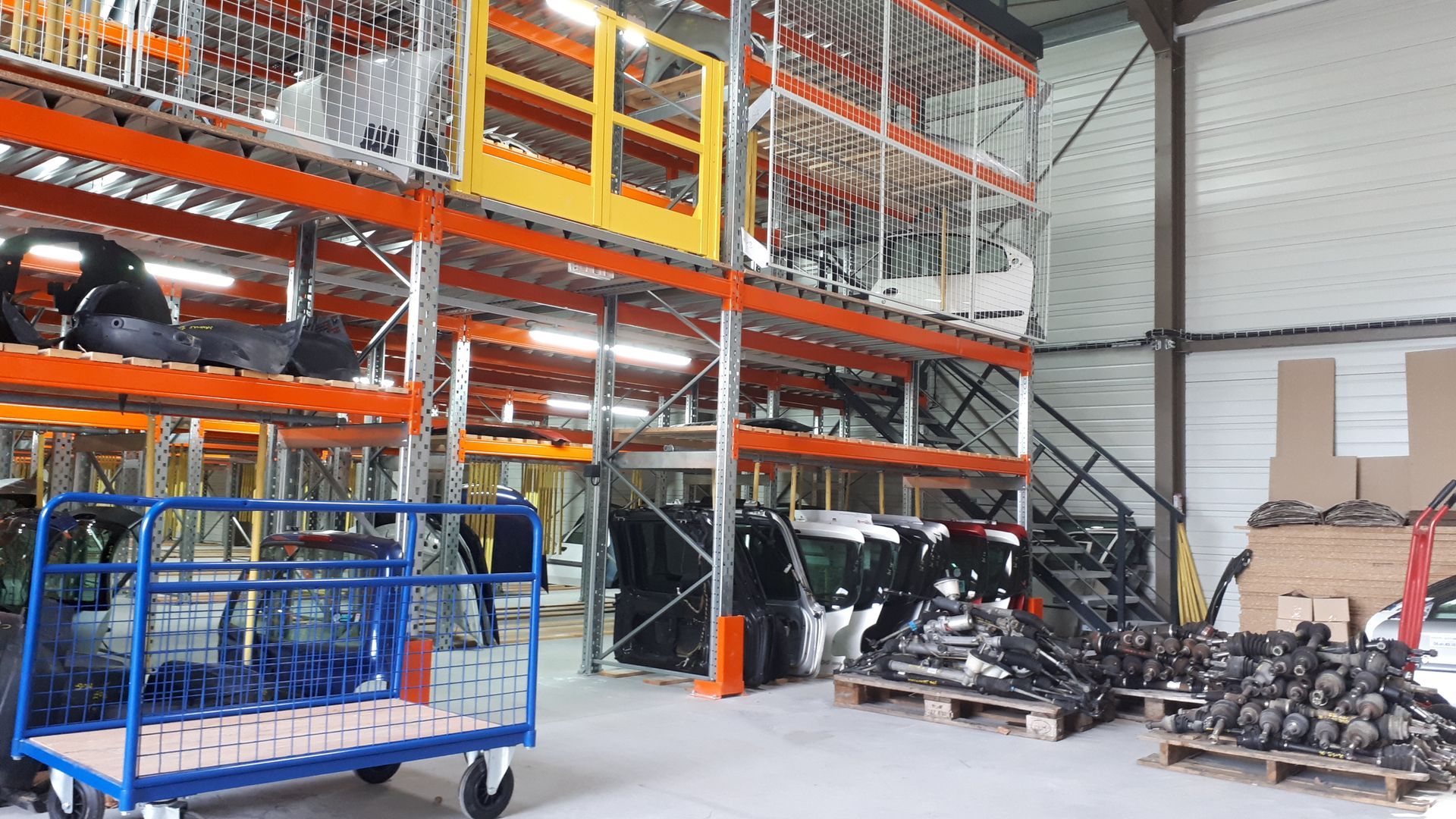 Intérieur d'un entrepôt avec des étagères à plusieurs niveaux contenant des pièces automobiles. Un chariot bleu se trouve au premier plan.