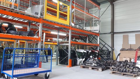 Intérieur d'un entrepôt avec des étagères à plusieurs niveaux contenant des pièces automobiles. Un chariot bleu se trouve au premier plan.