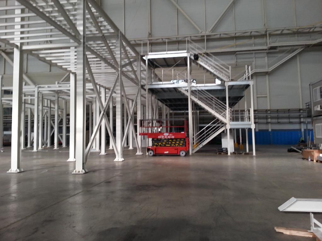 Grand espace industriel avec étagères métalliques blanches, escaliers et ascenseur rouge.