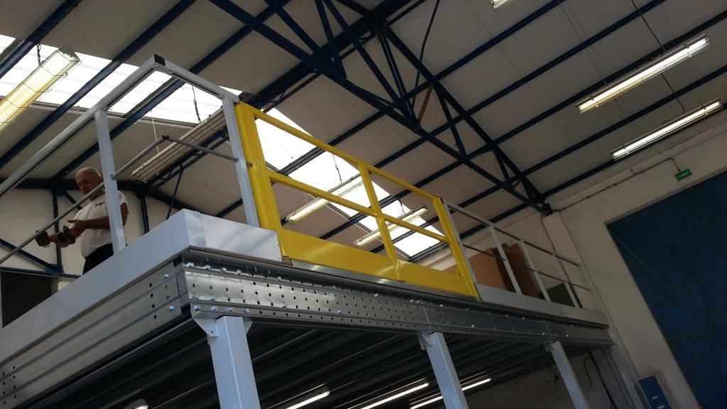 Un homme travaille sur une mezzanine équipée d'une barrière de sécurité jaune dans un entrepôt.