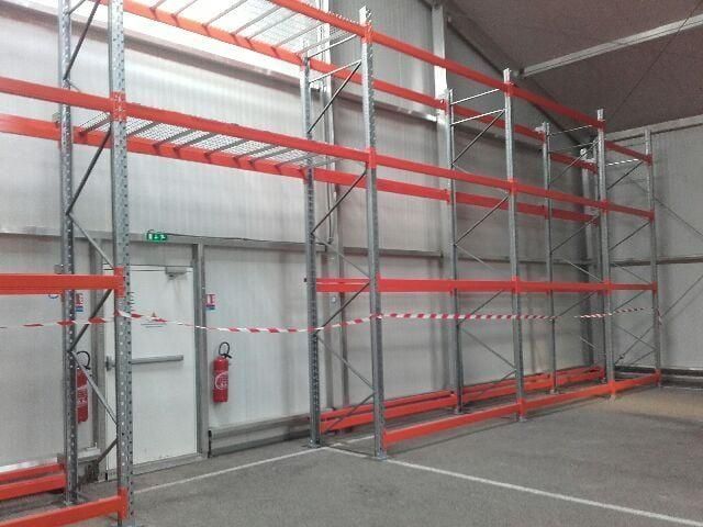 Intérieur d'entrepôt avec des étagères rouges et argentées le long d'un mur blanc, une porte fermée et un extincteur.