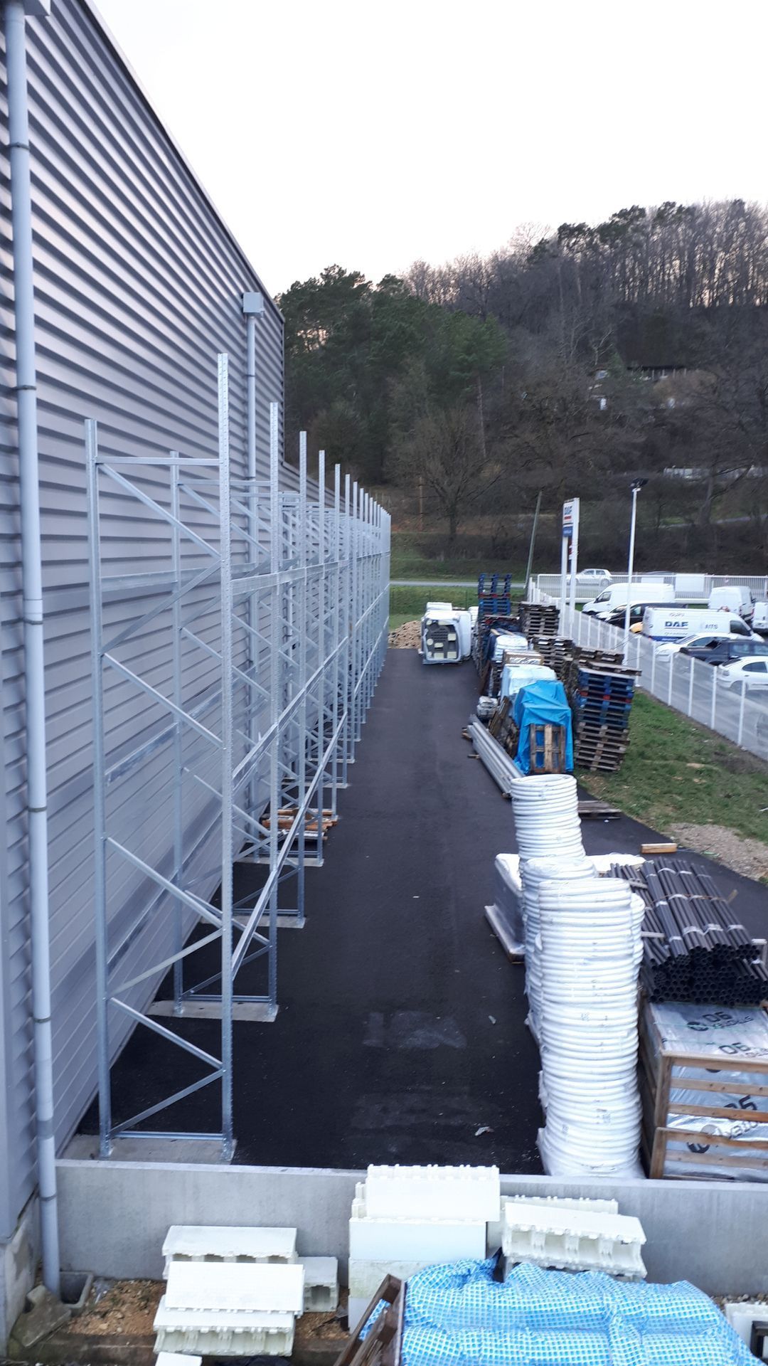 Des rayonnages métalliques jouxtent un grand bâtiment industriel, un chemin d'asphalte noir et des véhicules stationnés.