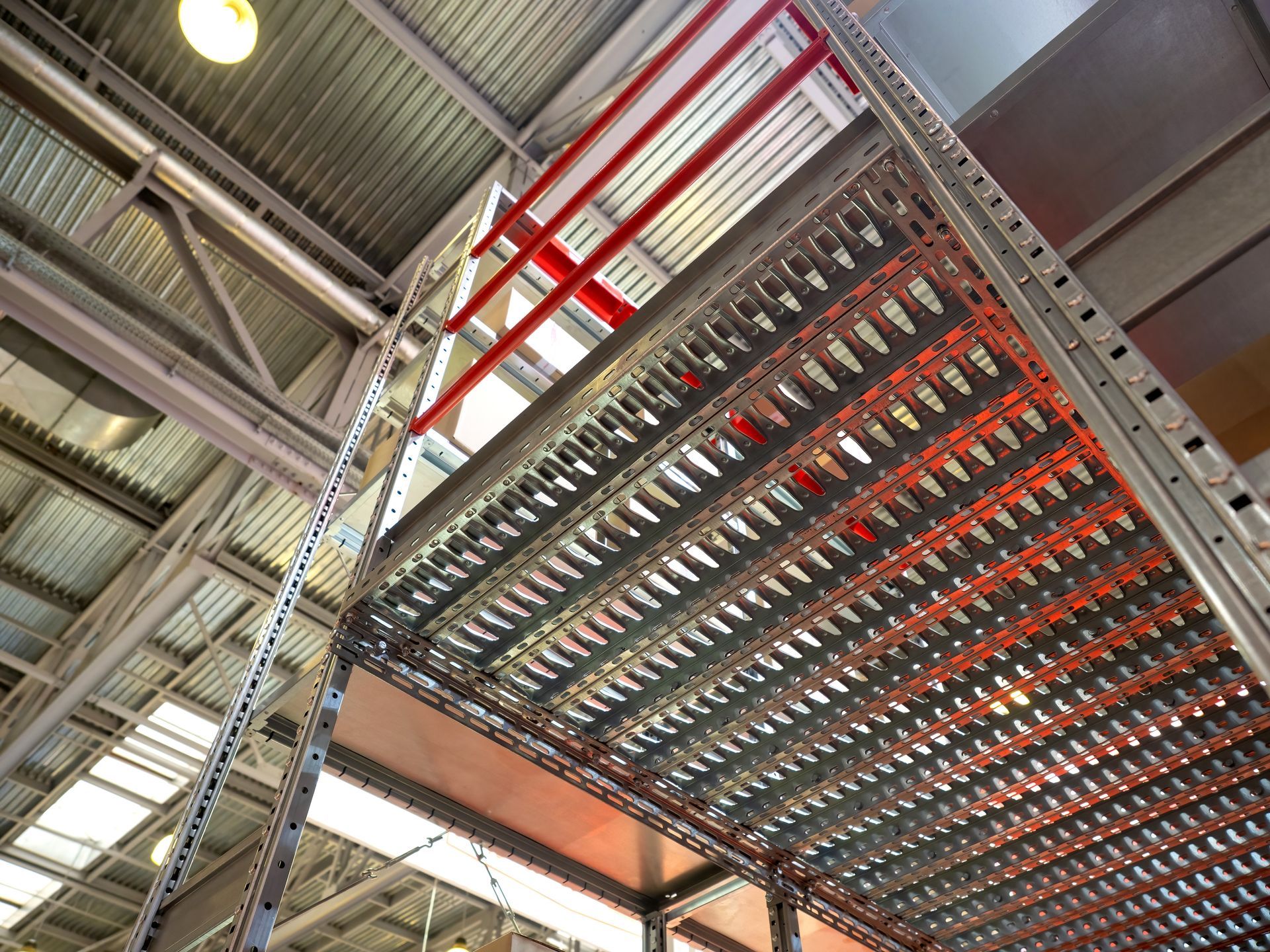 Rayonnages industriels en acier, avec étagères métalliques perforées, vus de dessous. Poutres de soutien rouges.