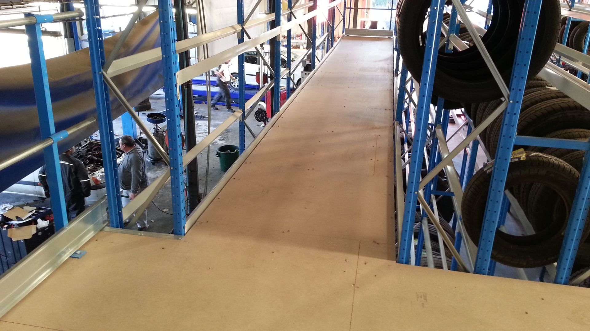 Rampe dans un entrepôt, flanquée d'étagères métalliques avec des pneus à droite. Des personnes en arrière-plan.