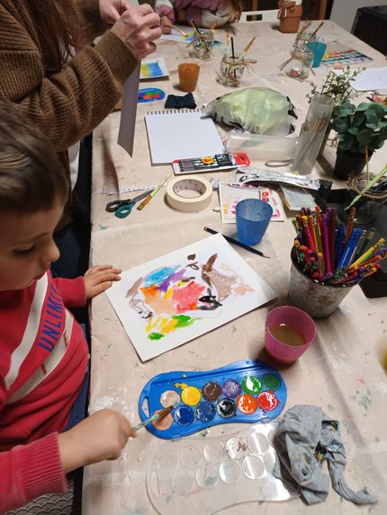Un niño pinta con acuarelas sobre un papel en una mesa desordenada con materiales de arte.