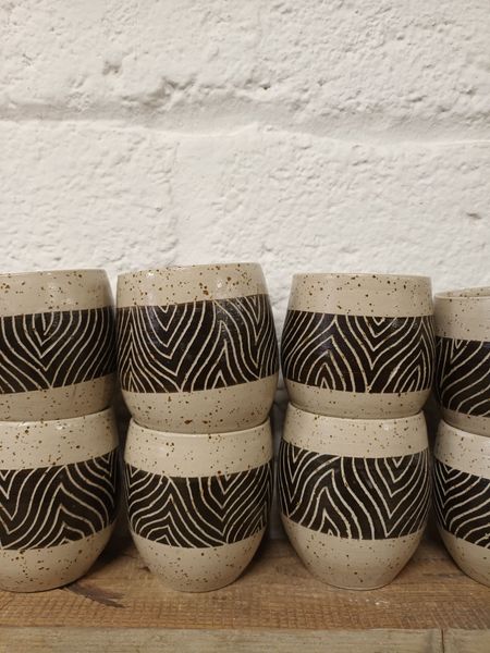 Una hilera de tazas de cerámica beige moteadas con bandas oscuras en zigzag, dispuestas en un estante de madera contra una pared blanca.