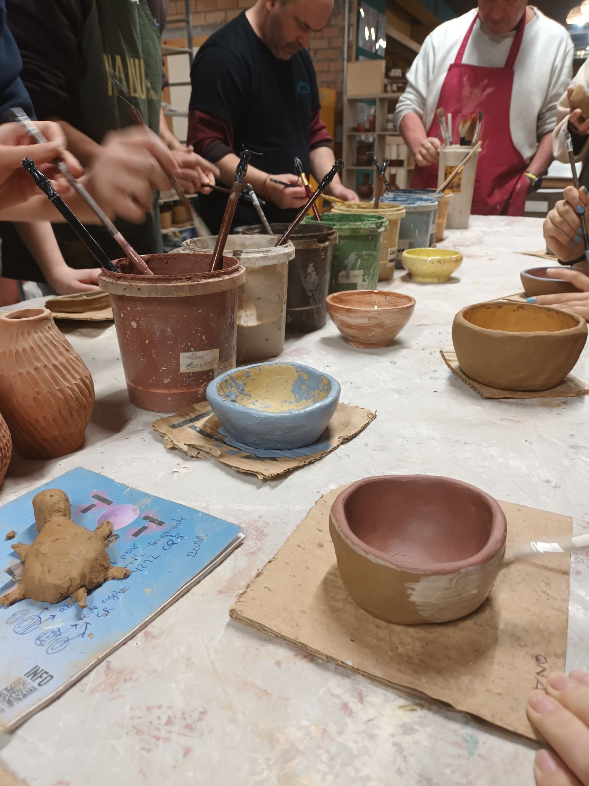 En un taller de cerámica, la gente trabaja sentada en una mesa, pintando cuencos de cerámica y creando figuras de arcilla con pinceles y esmalte.