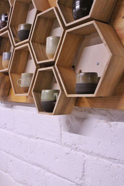 Tazas de cerámica hechas a mano expuestas en estantes hexagonales de madera sobre una pared de ladrillo blanco.