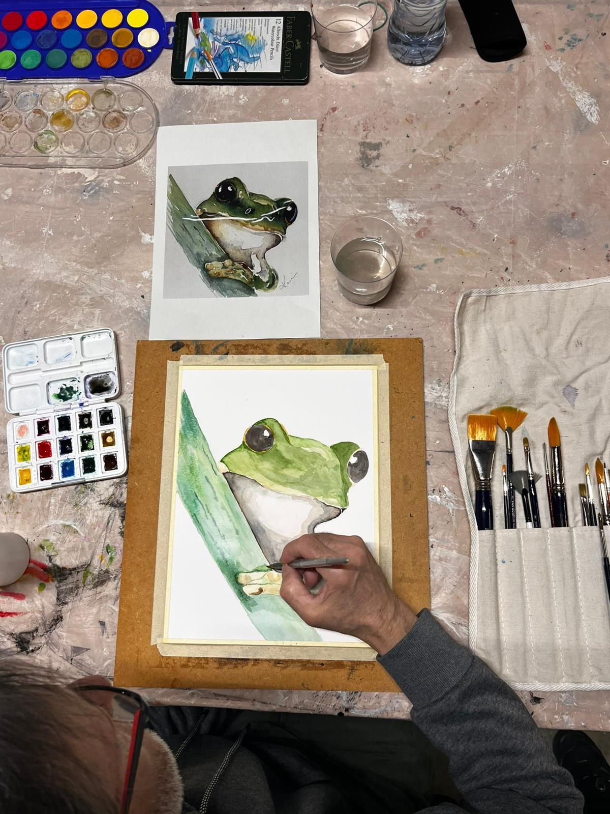 Un artista pinta una rana en acuarela sobre un escritorio lleno de pinceles, una foto de referencia y paletas de pintura.