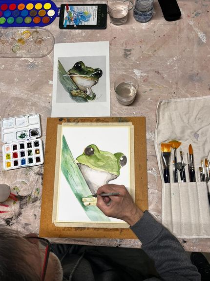 Un artista pinta una rana en acuarela sobre un escritorio lleno de pinceles, una foto de referencia y paletas de pintura.
