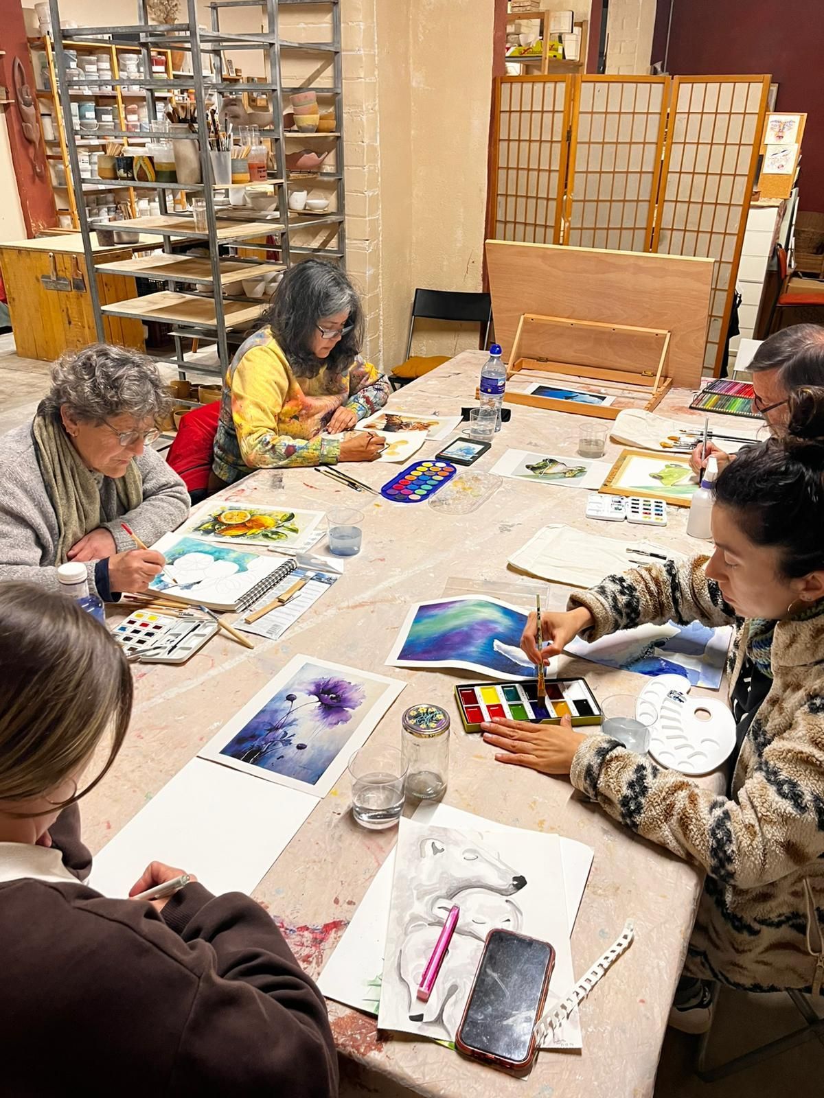 Un grupo de personas se sienta alrededor de una mesa larga en un estudio, concentradas en pintar acuarelas sobre papel.