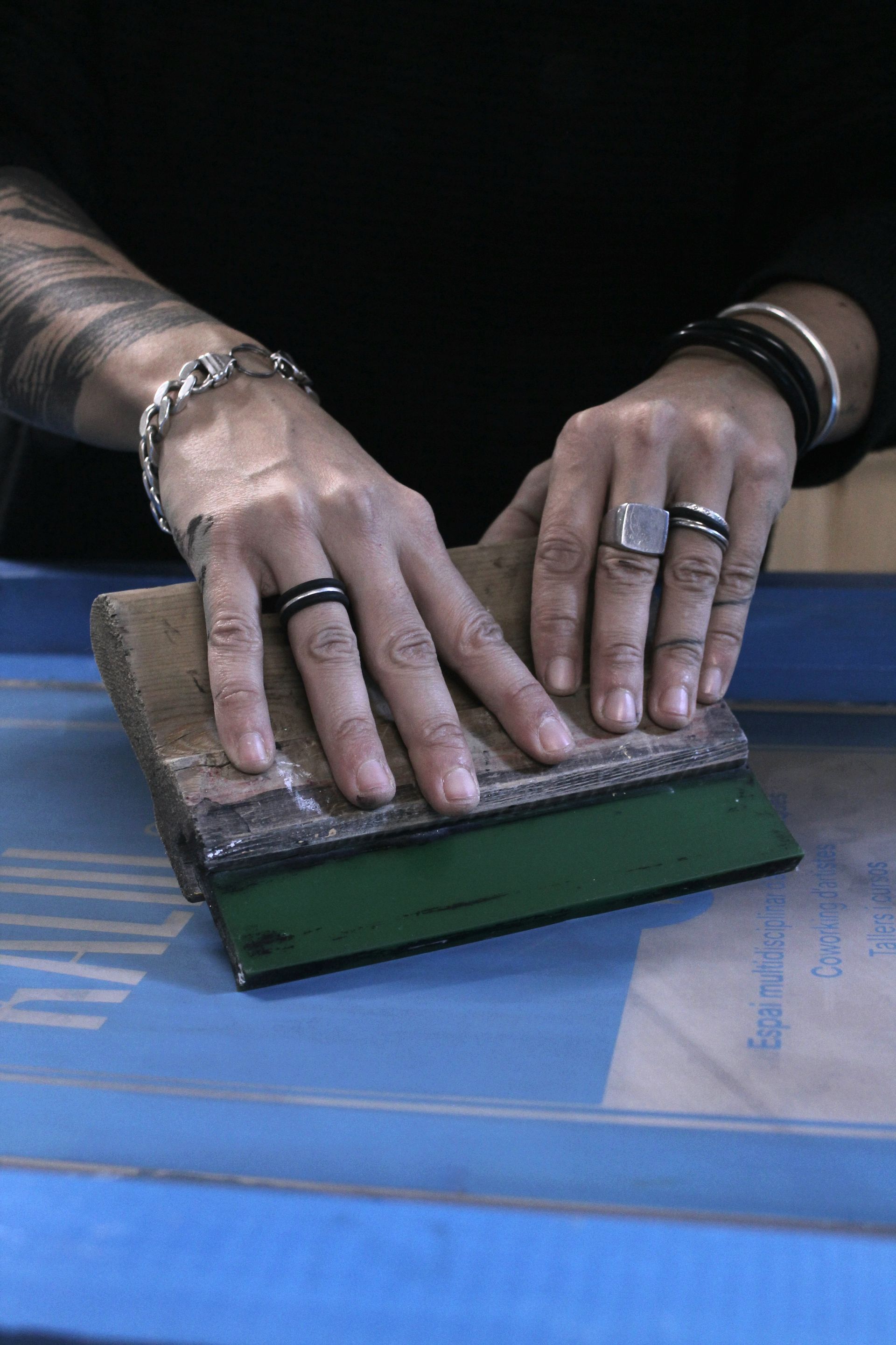 Unas manos adornadas con anillos y pulseras presionan una espátula verde sobre una malla azul de serigrafía.