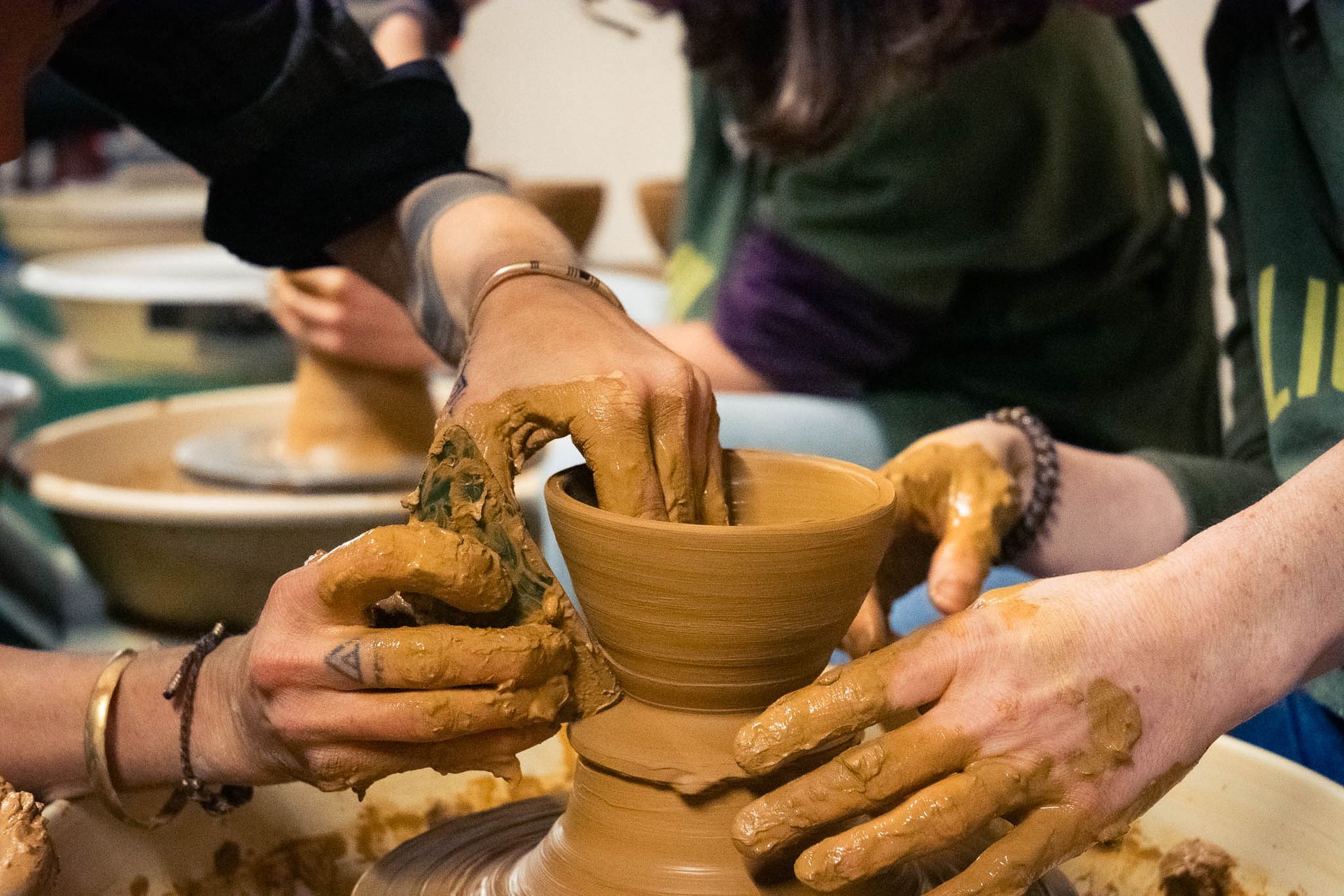 Primer plano de unas manos cubiertas de arcilla húmeda dando forma a una vasija en un torno de alfarero giratorio, con otras personas trabajando al fondo.