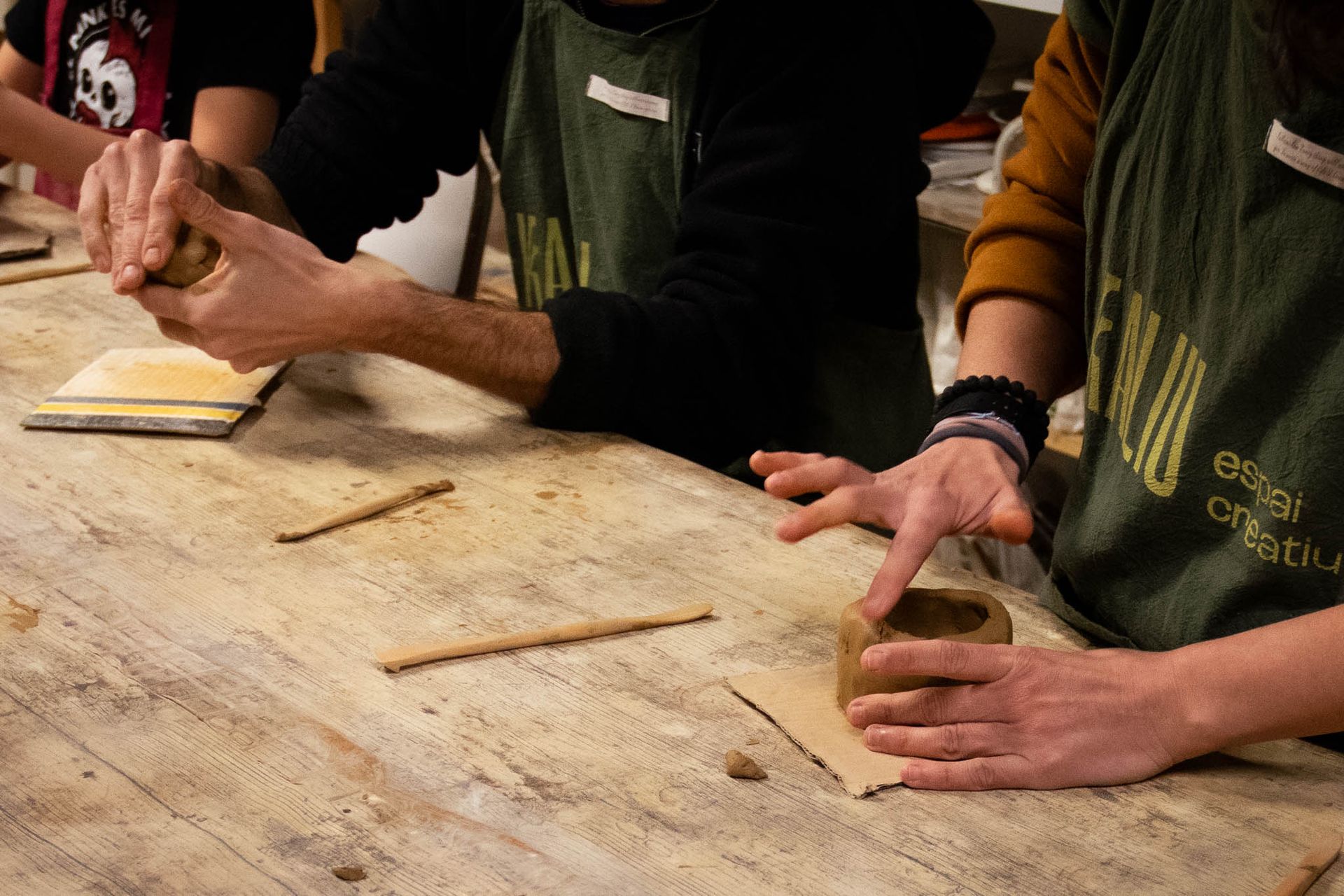 Gent donant forma a bols i tasses d'argila sobre una taula de taller amb eines i argila humida.