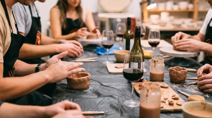 Durante una clase de cerámica, la gente se sienta a una mesa con delantales, trabaja con arcilla y bebe vino.
