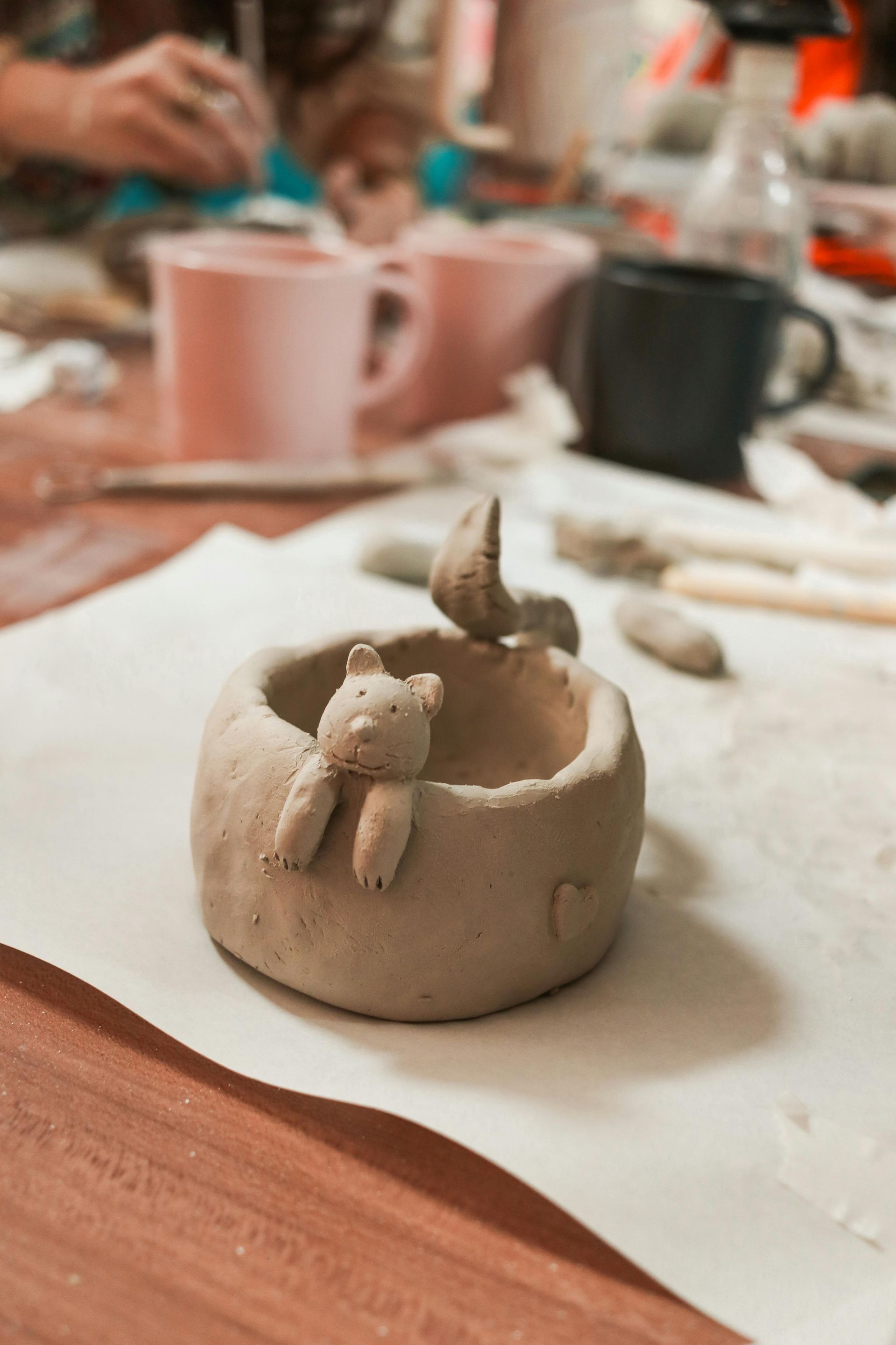 Un pequeño cuenco de arcilla esculpido a mano con la figura de un oso apoyada en el borde, sobre papel blanco en un taller de cerámica.