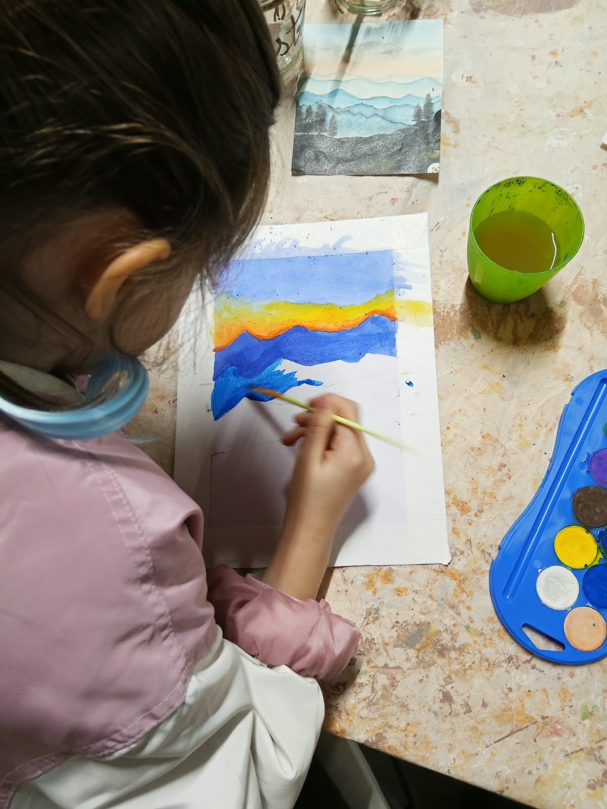 Gent pintant amb aquarel·les a una taula, amb pinzells, pintures i un got d'aigua.