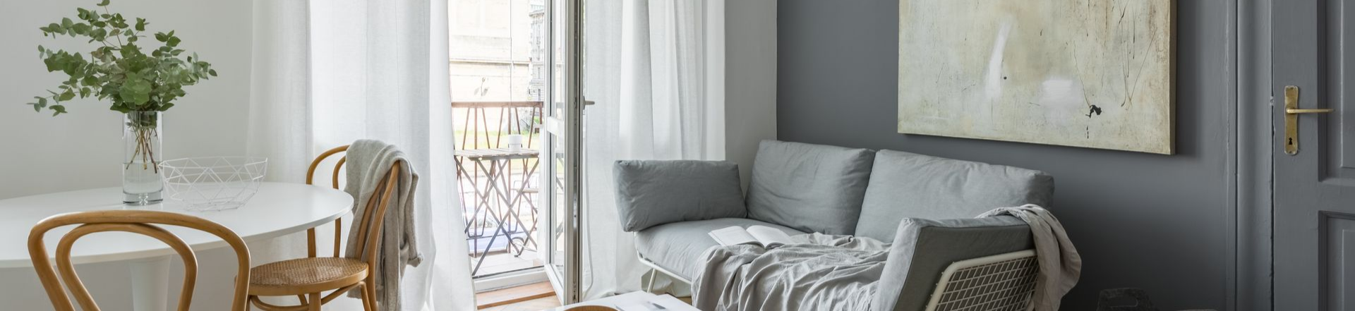 Salon avec un petit canapé, une table et des chaises, et un balcon. Murs gris, meubles clairs.