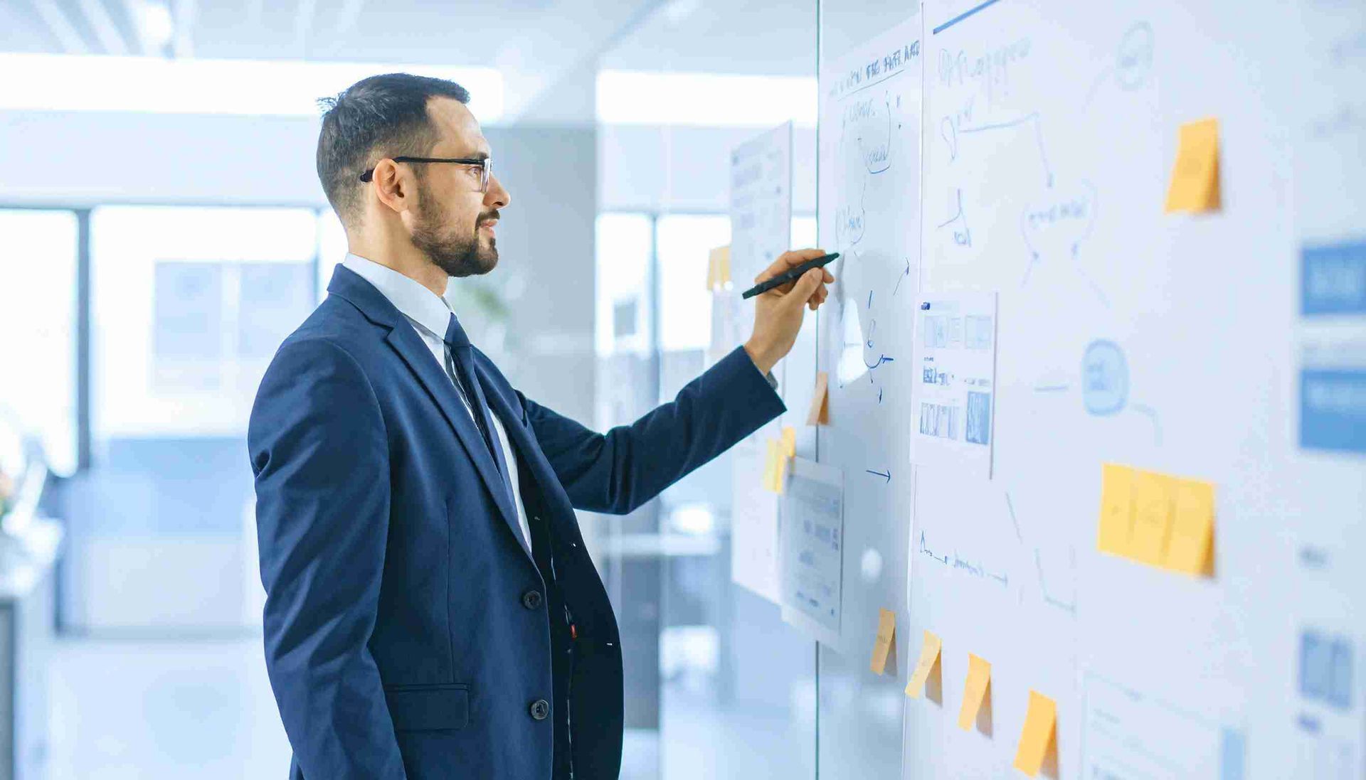 Hombre con traje y gafas escribiendo en una pizarra con diagramas y notas adhesivas en un entorno de oficina.
