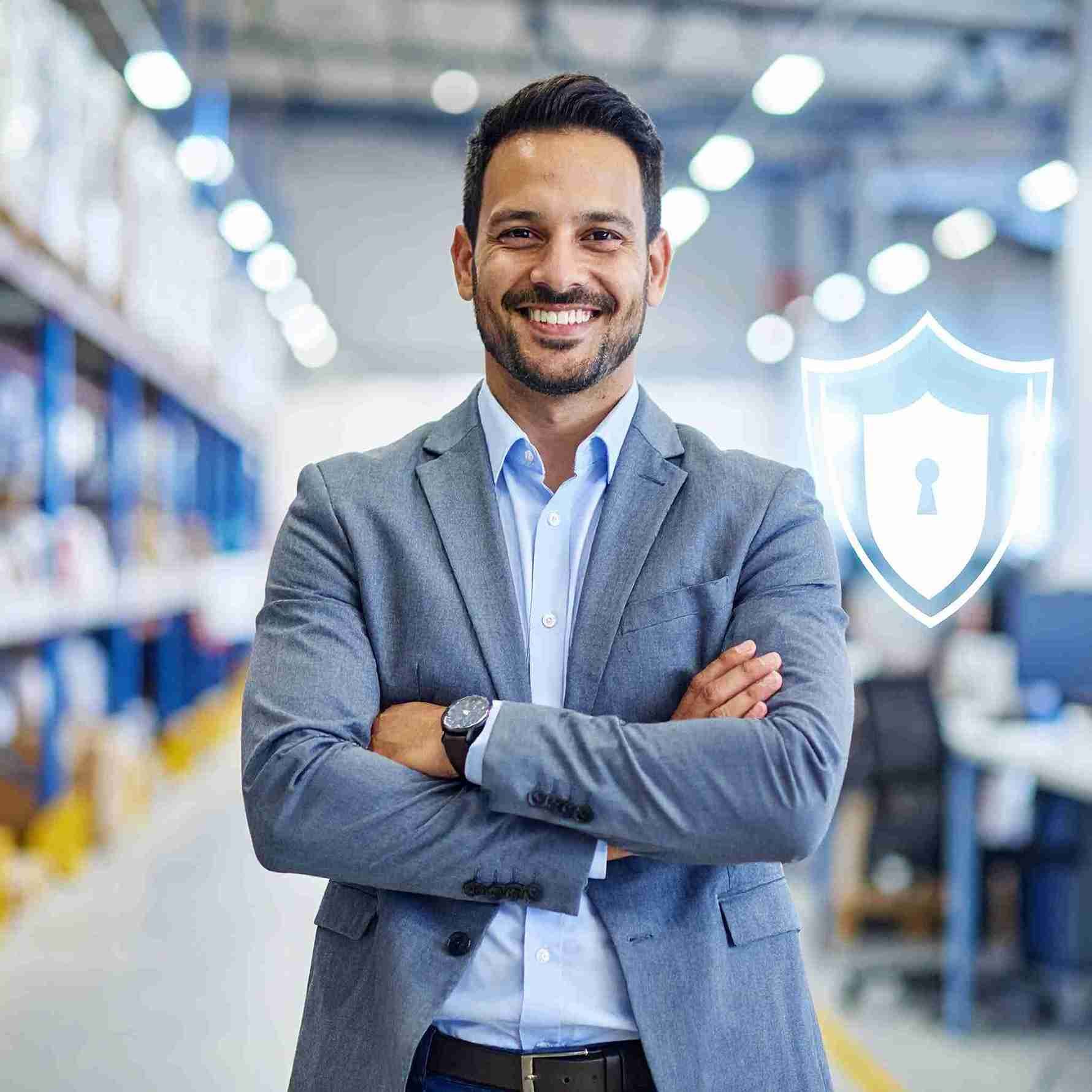 Un hombre sonriente con traje gris y brazos cruzados, de pie en un almacén. Junto a él hay un icono de escudo de seguridad.