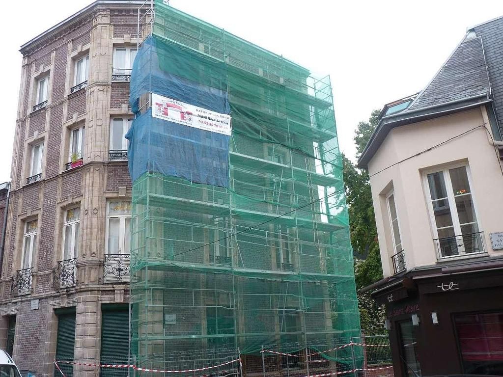 Un grand bâtiment en briques est recouvert d'échafaudages et de grillages verts, situé entre deux autres maisons de ville.