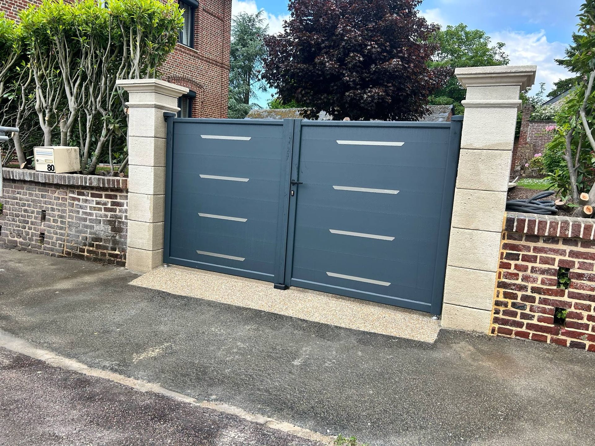 Un portail double gris foncé orné de bandes horizontales argentées se trouve entre deux piliers de pierre, reliés par un muret de briques.