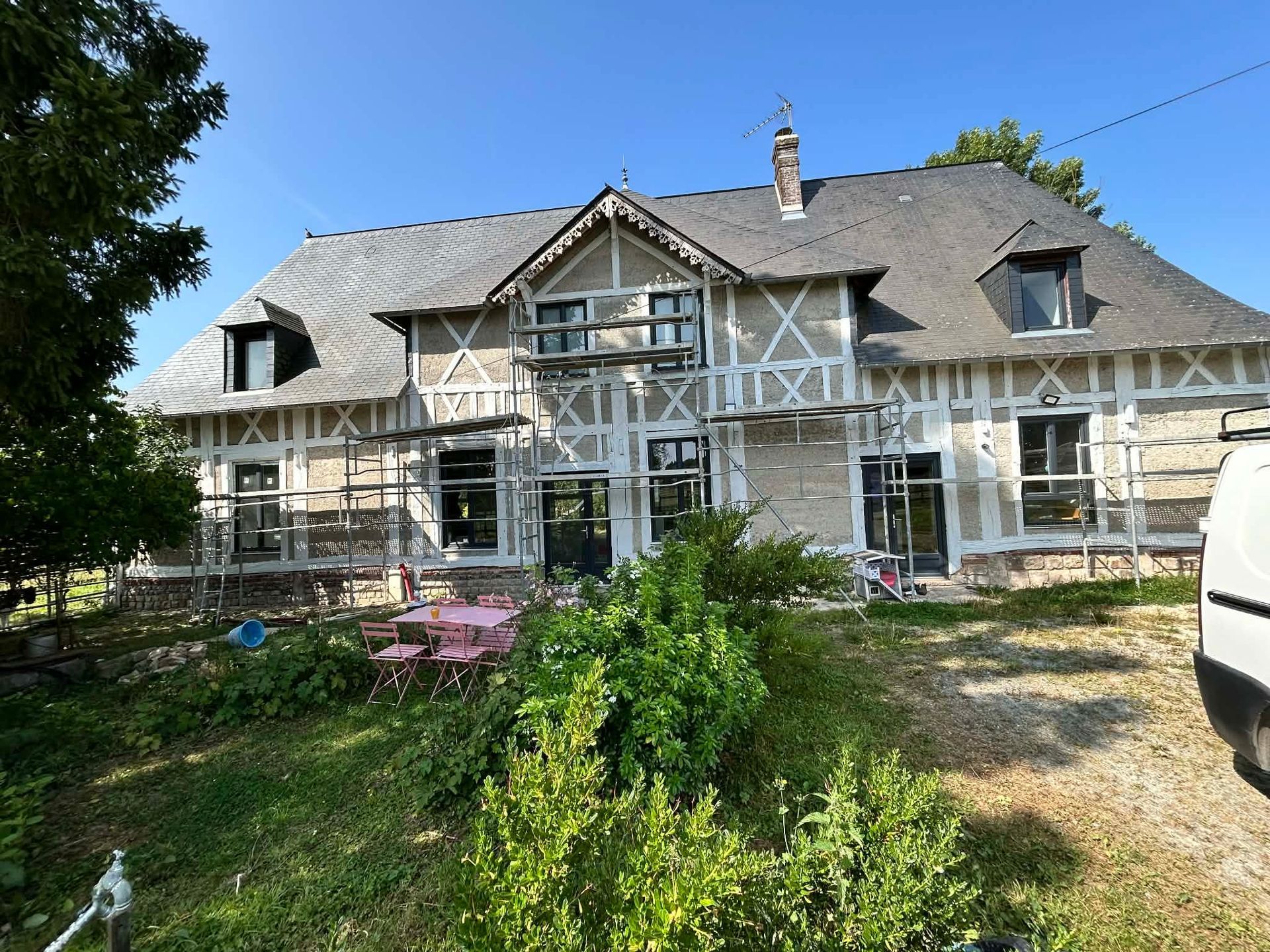 Une maison à colombages avec un toit en ardoise grise, en cours de rénovation extérieure avec des échafaudages dans un jardin ensoleillé.