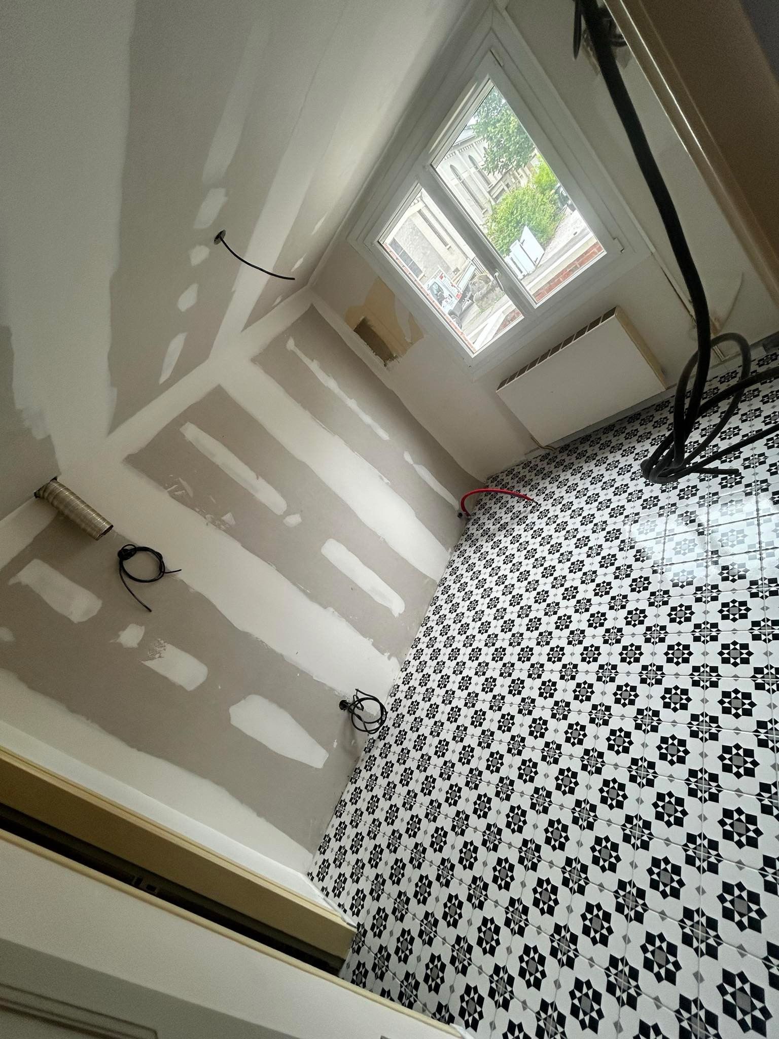 Une pièce en rénovation avec des murs en plaques de plâtre, un sol carrelé à motifs noirs et blancs, une fenêtre et des fils électriques apparents.