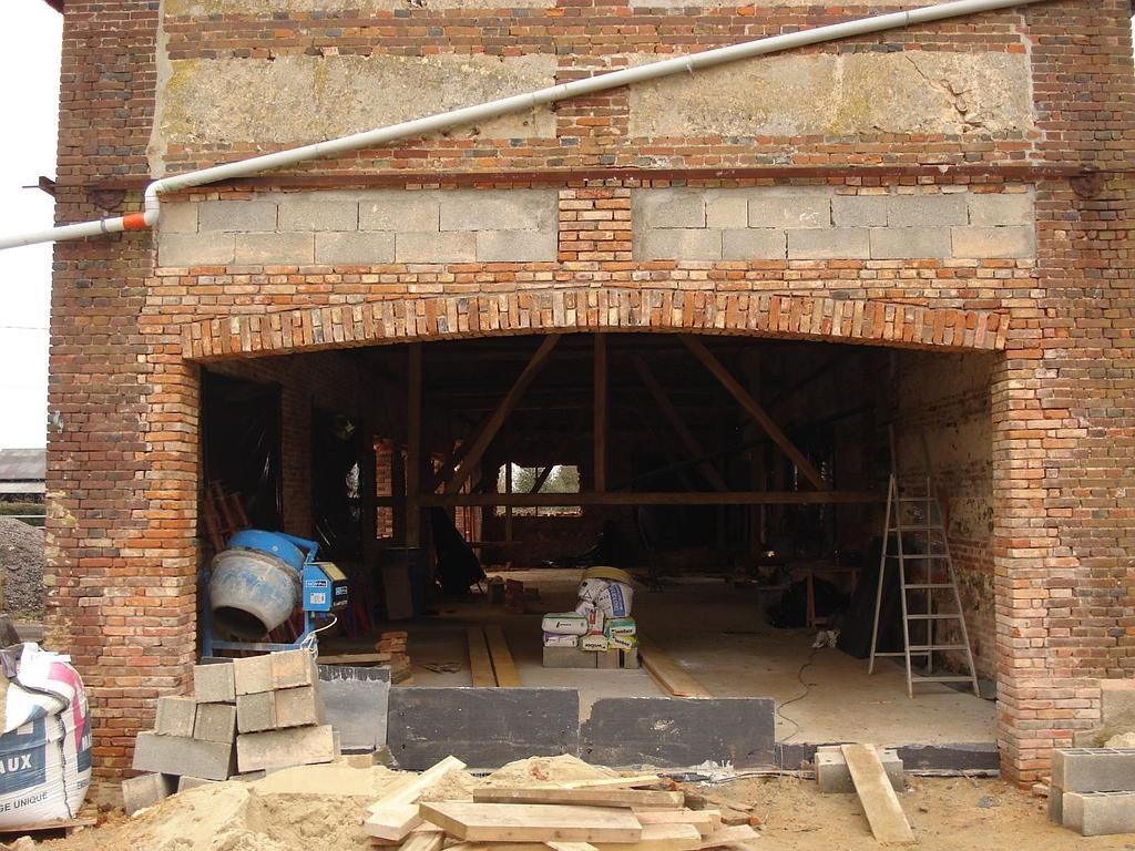 Un chantier de construction devant un bâtiment en briques avec une grande porte cintrée ouverte, une bétonnière et des matériaux de construction.