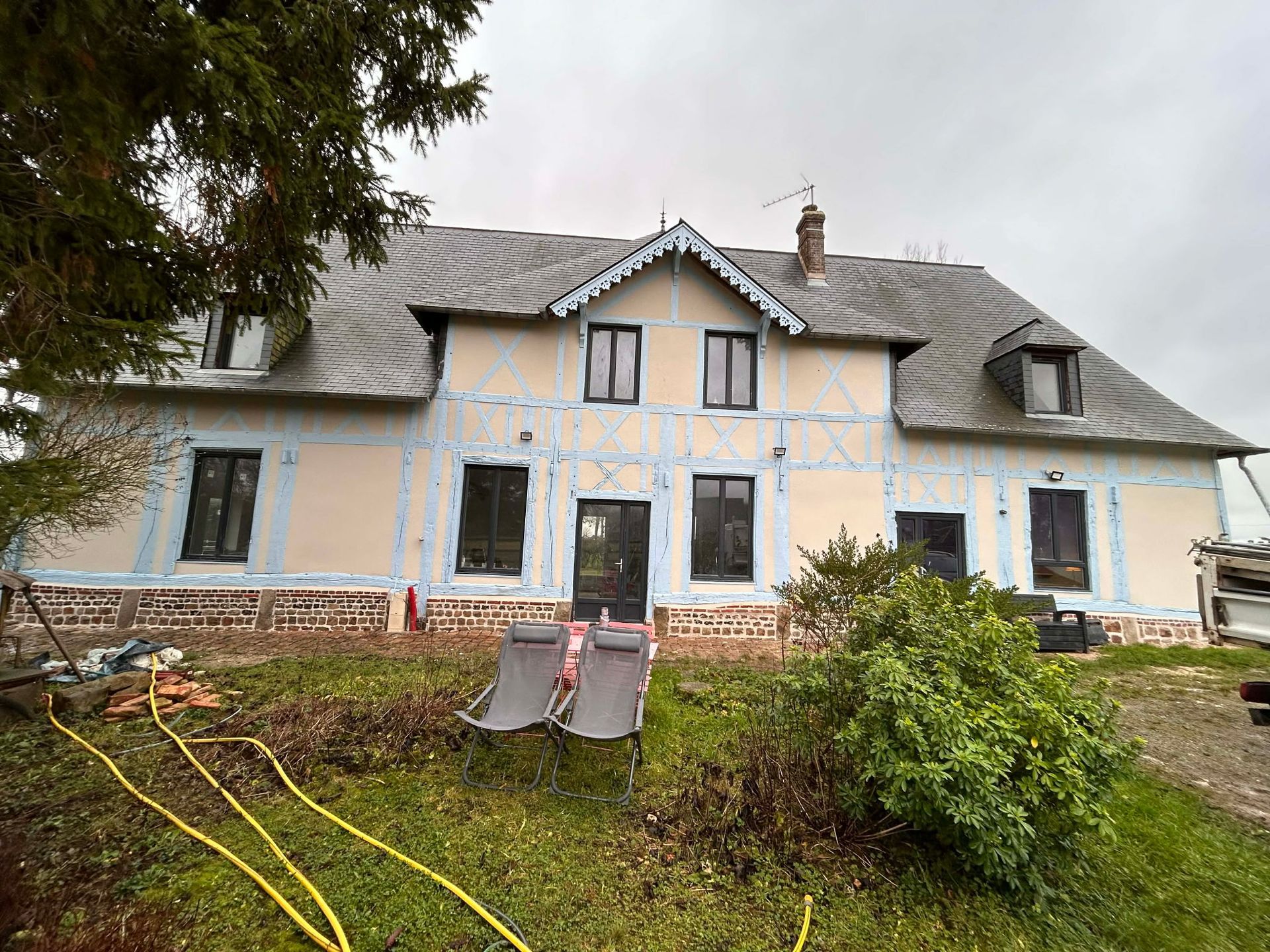 Une maison à colombages de deux étages avec un toit en bardeaux gris, située derrière une pelouse avec deux chaises de patio et un tuyau d'arrosage jaune.