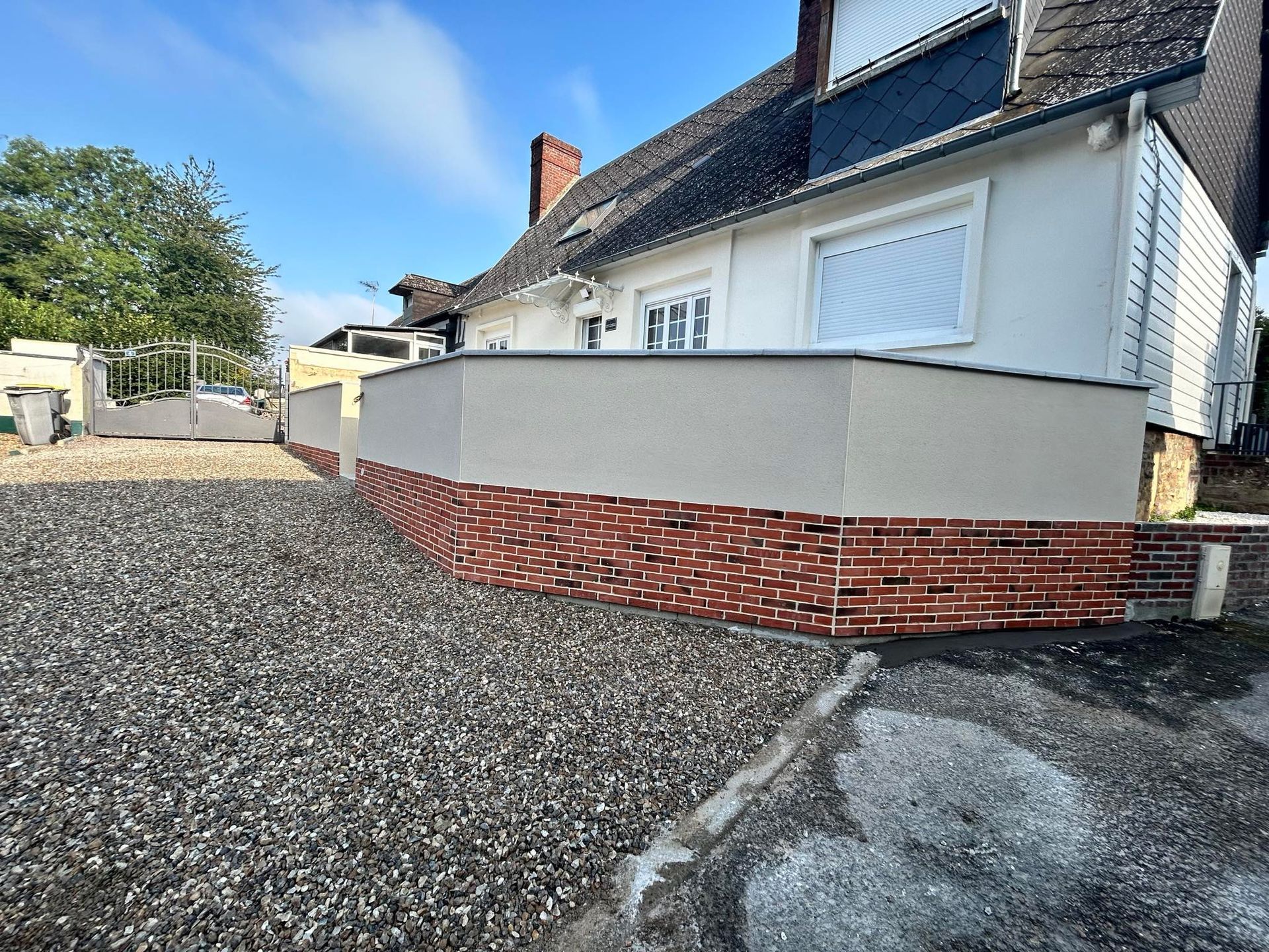 Une maison blanche avec un mur enduit clair neuf et un soubassement en briques rouges décoratives, située à côté d'une cour gravillonnée.