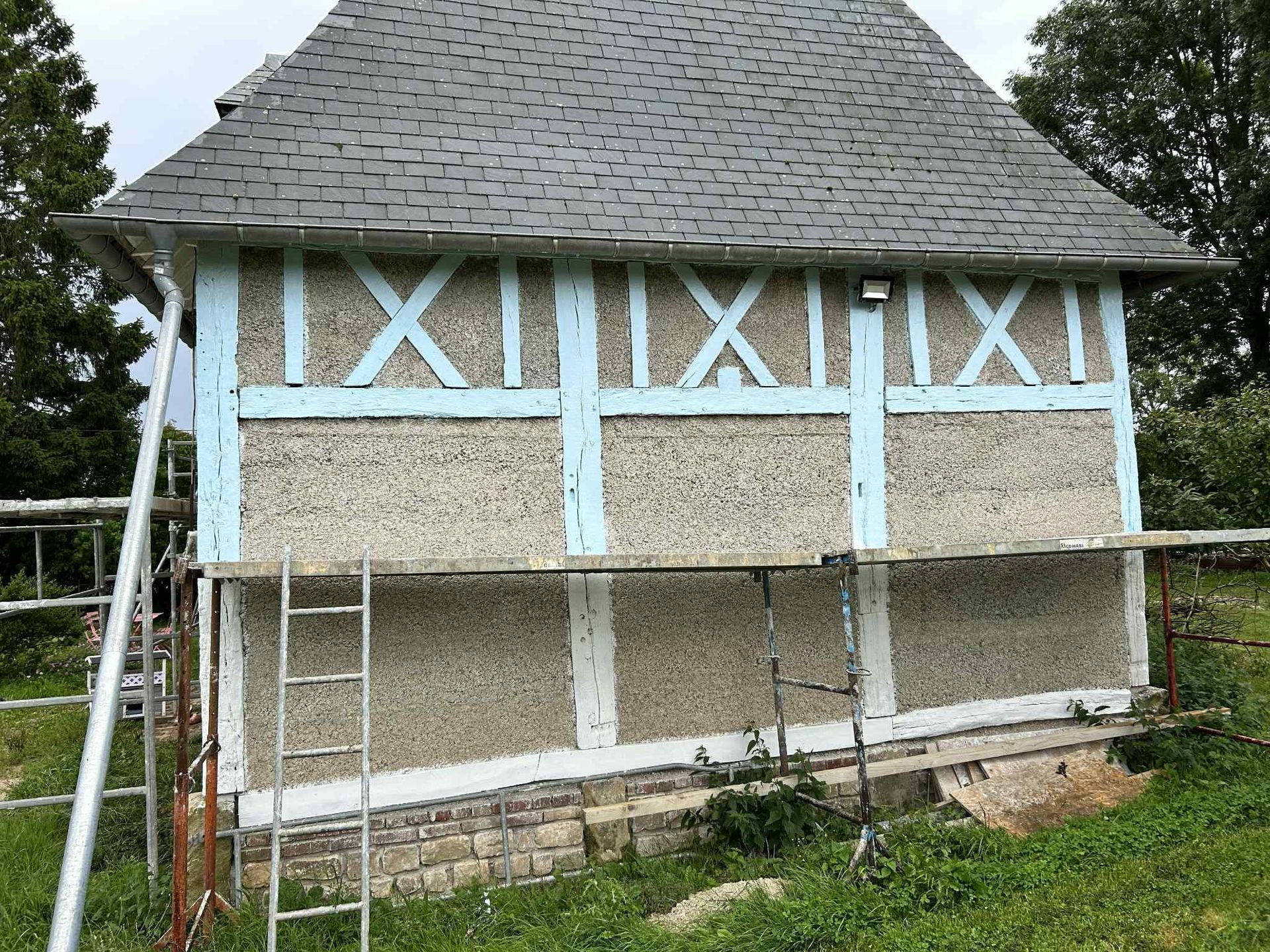 Vue latérale d'une maison à colombages en cours de rénovation, avec ses poutres en bois bleu clair et ses murs texturés de gravier.