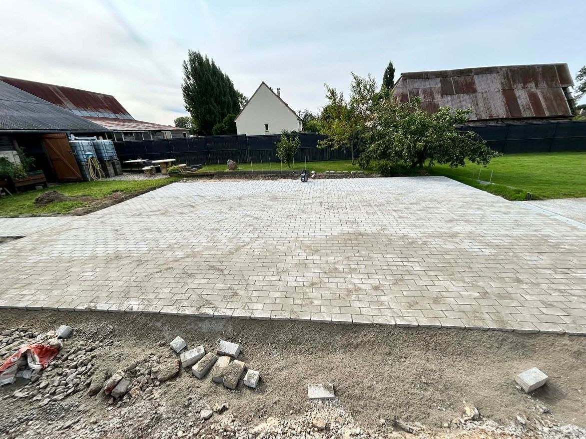 Une terrasse en pavés de pierre gris clair, récemment posée, se trouve dans un jardin avec une clôture sombre, des arbres et des maisons en arrière-plan.