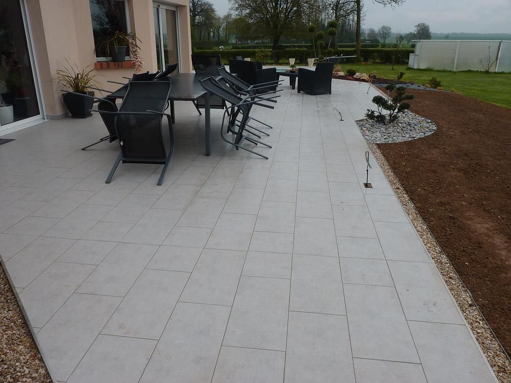 Une terrasse carrelée gris clair avec un ensemble de salle à manger noir et des chaises longues, attenante à l'extérieur d'une maison et à un jardin.