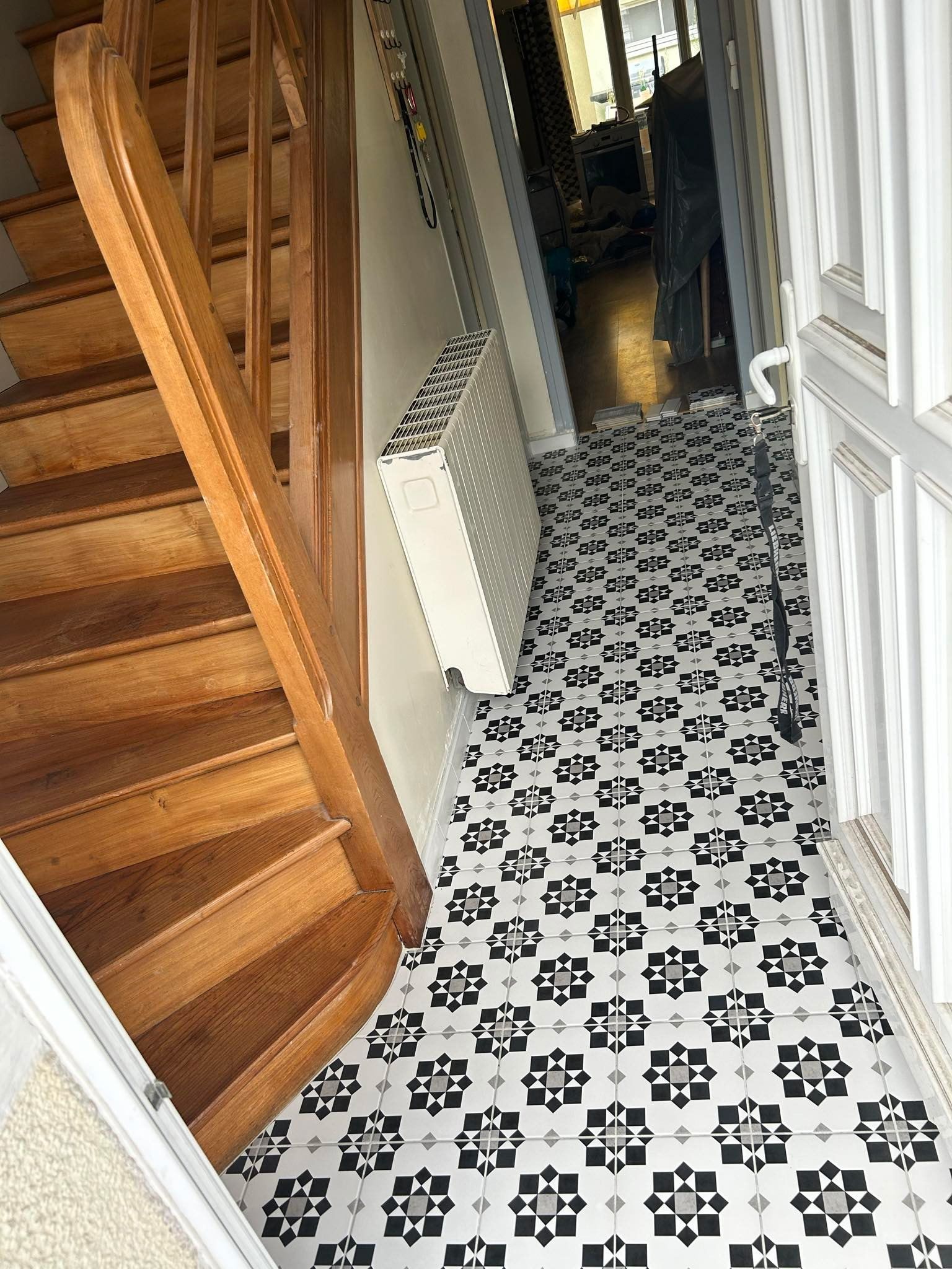 Un escalier en bois jouxte un couloir avec un carrelage au sol à motifs noirs et blancs et un radiateur mural.