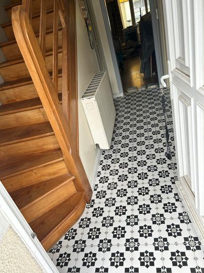 Une entrée avec un escalier en bois sur la gauche, un radiateur mural et un sol carrelé à motifs noirs et blancs.