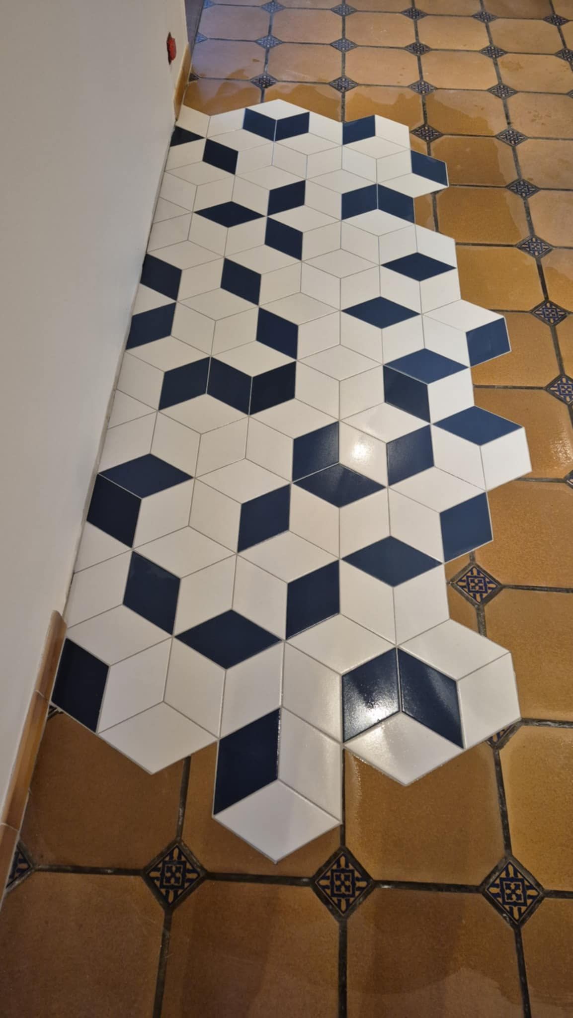 Une section de carreaux blancs et bleu foncé en forme de losange, disposés en motif cubique 3D, posée sur un sol carrelé beige existant.