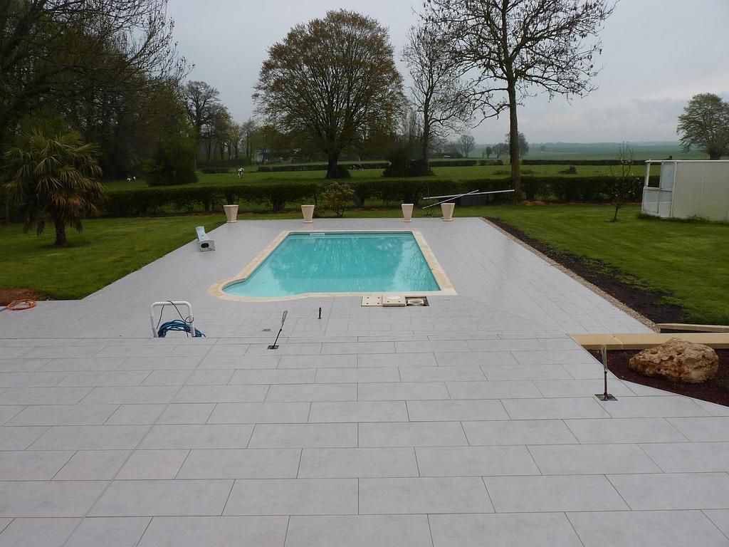 Une piscine rectangulaire entourée d'une terrasse carrelée gris clair, le tout sur une pelouse verdoyante avec des arbres en arrière-plan.