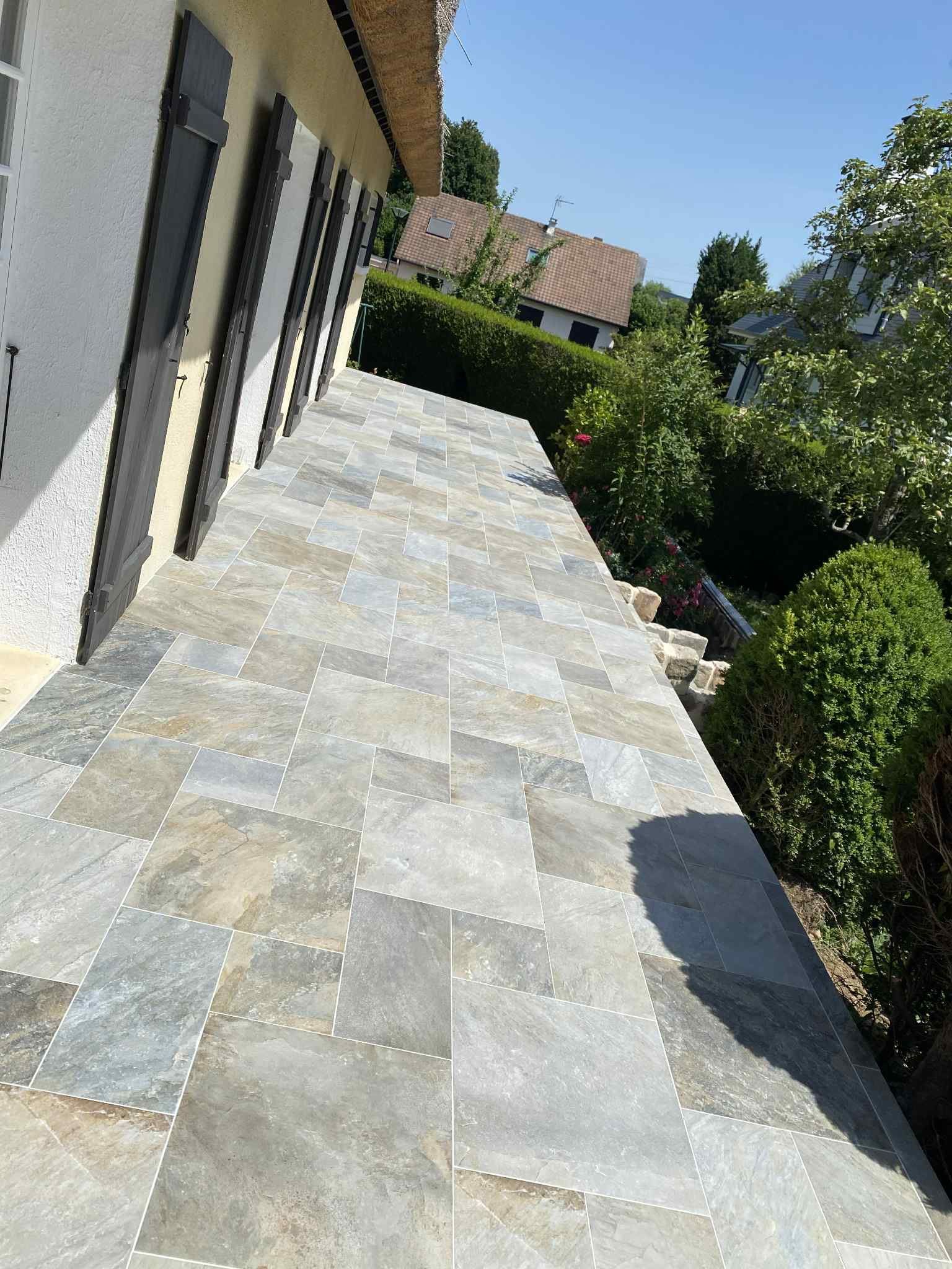 Une terrasse en pierre longe le côté d'une maison aux tons clairs et aux volets foncés, bordée d'une haie et d'arbres.