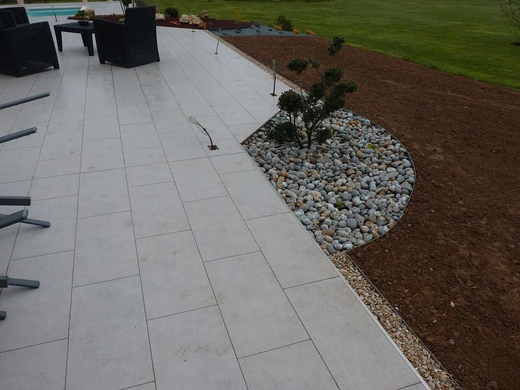 Une terrasse carrelée gris clair jouxte un parterre de jardin incurvé orné de galets décoratifs et de paillis, à côté d'une pelouse.