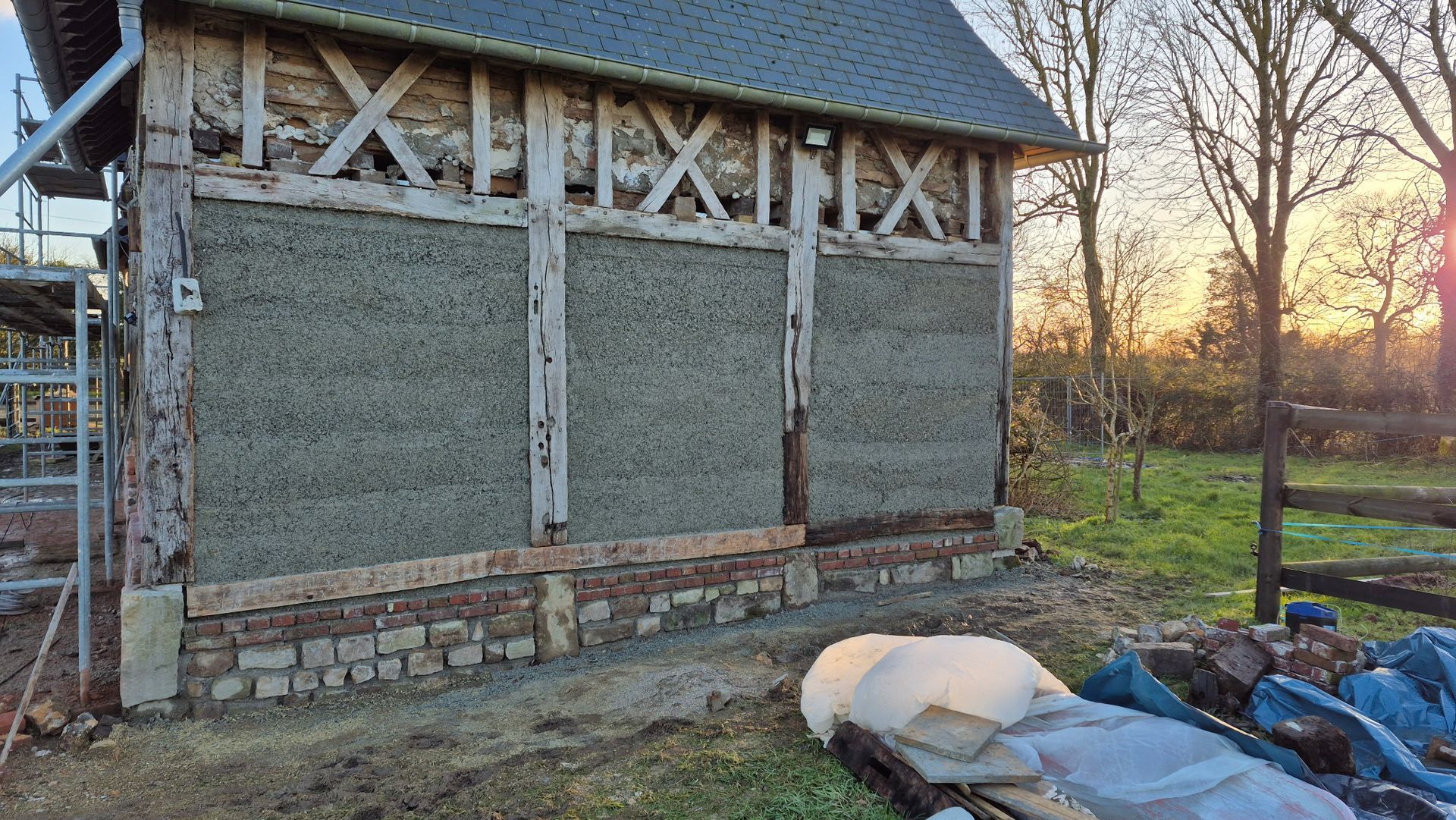 Un bâtiment à ossature bois en cours de rénovation, avec charpente apparente, panneaux de remplissage et fondations en pierre et en brique.